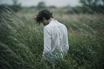 Medium-long shot from behind, slightly angled left, standing centered in tall windswept wild grass. Slender build, crisp long-sleeved white shirt