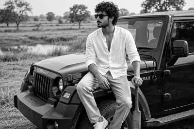 A cinematic, black and white, outdoor portrait of a stylish young man sitting on the front fender of a black Thar SUV in a rural landscape, captured
