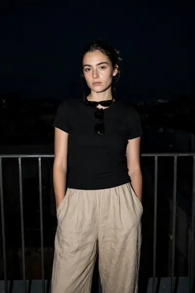 Dark Night portrait, woman standing upright against railing, calm neutral face. Wearing a simple fitted black top with loose linen pants. Clean