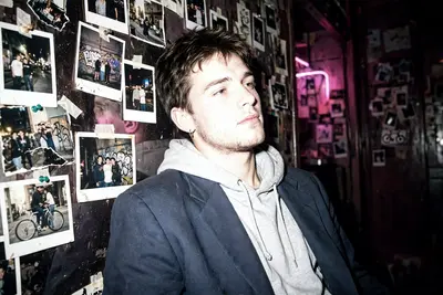 A candid 90s flash photo of a young man with the same face and relaxed posture. Wearing a casual hoodie layered under a tailored blazer. Background
