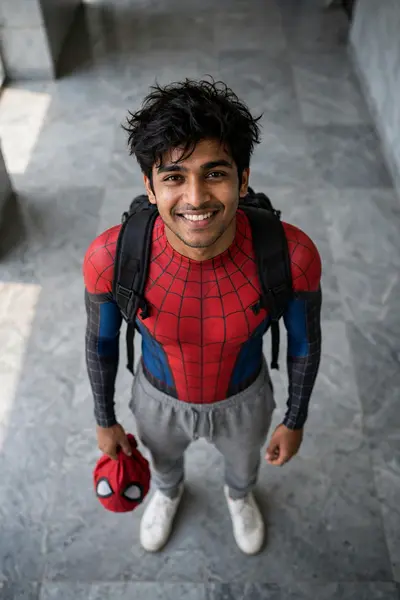high-angle full-body shot of a young South Asian man looking straight up at the camera with a warm, confident smile, standing alone on a grey