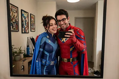high-angle full-body shot of a South Asian couple looking straight up at the camera with warm, genuine smiles, standing side by side on a grey