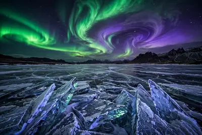 Vivid green and violet aurora borealis swirling over a frozen Icelandic lake, jagged ice shards glowing in the foreground. Night landscape