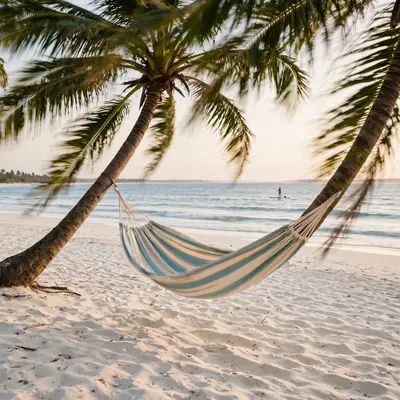 A striped hammock swaying between leaning coconut palms over powdery white sand, gentle waves and a paddleboarder in the distance. Tropical escape