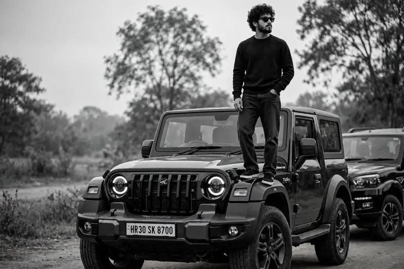 A cinematic, black and white editorial photograph of a stylish young man with curly hair, beard and mustache, wearing black sunglasses, standing