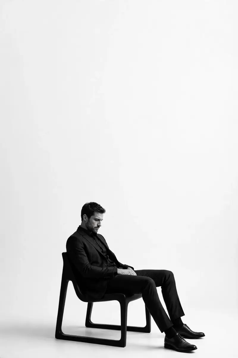 A high-contrast black and white editorial studio portrait of a man sitting in a sleek, minimalist chair, positioned in the lower third of the frame
