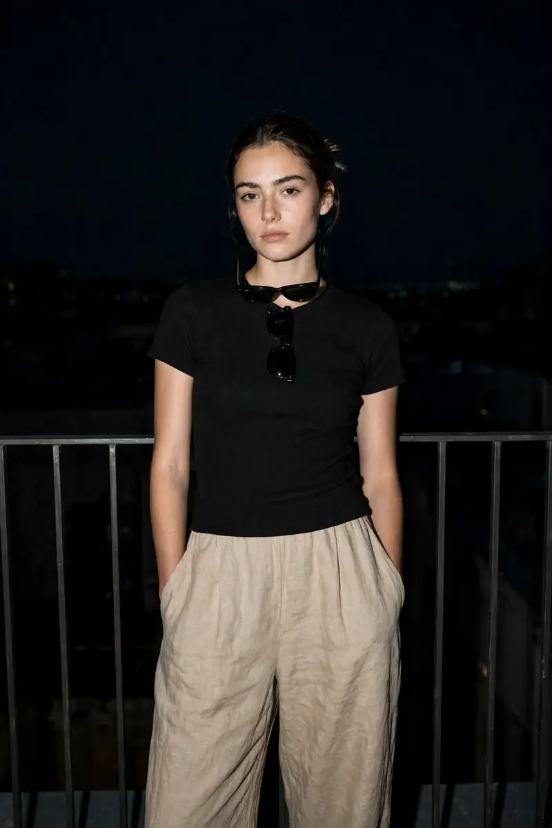Dark Night portrait, woman standing upright against railing, calm neutral face. Wearing a simple fitted black top with loose linen pants. Clean