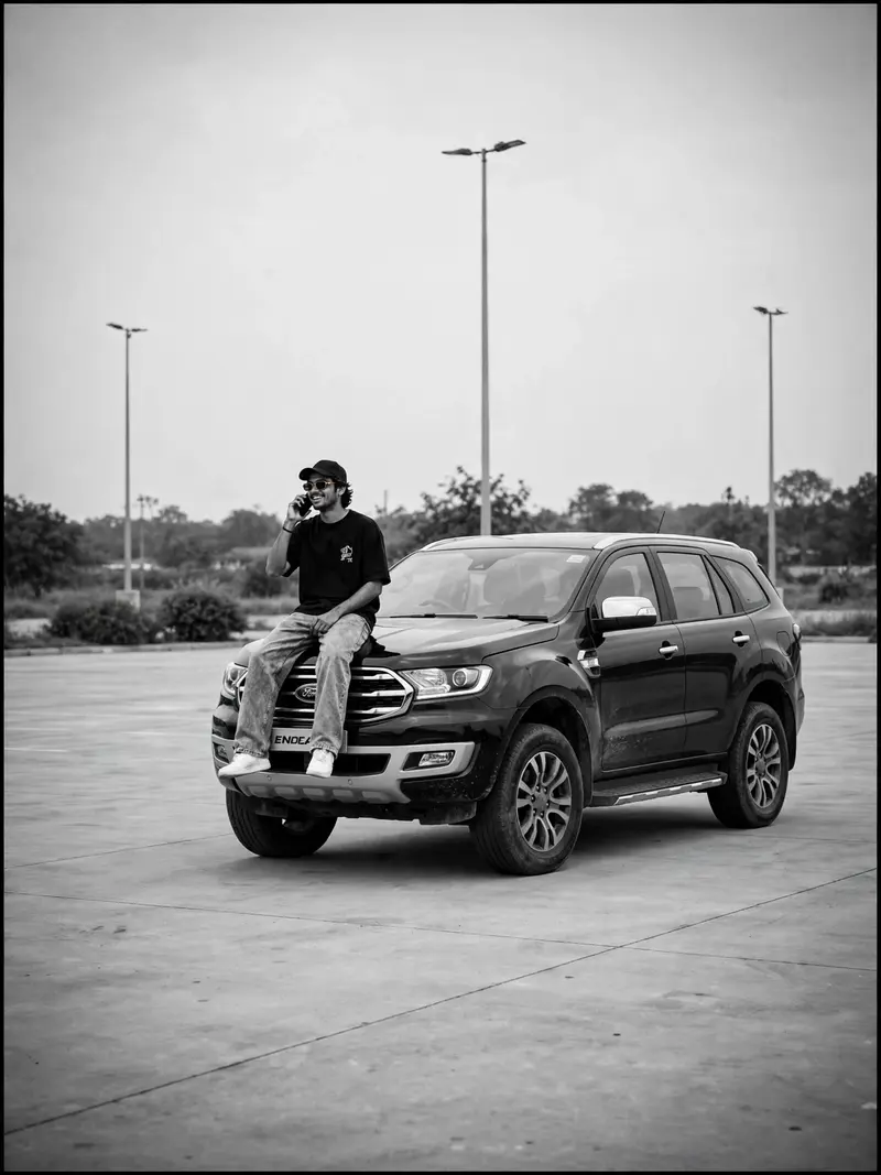 Black and white photo of a young South Asian man in his mid-20s sitting on the hood of a black Ford Endeavour SUV, front 3/4 low-angle view, the man