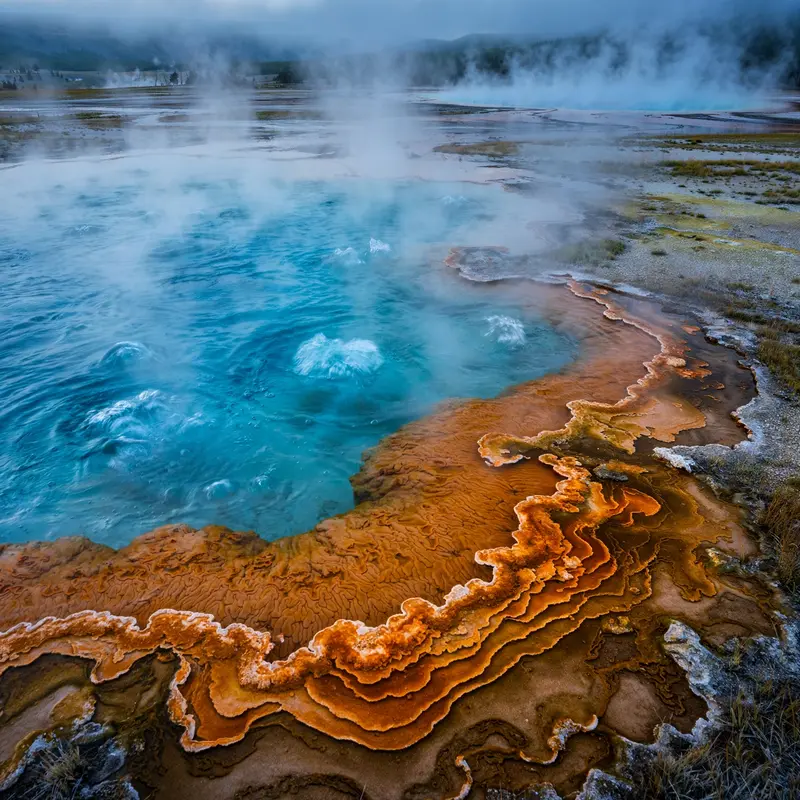 Bubbling turquoise hot springs and orange bacterial mats in Yellowstone's geothermal basin, steam curling in cold morning air. Geothermal