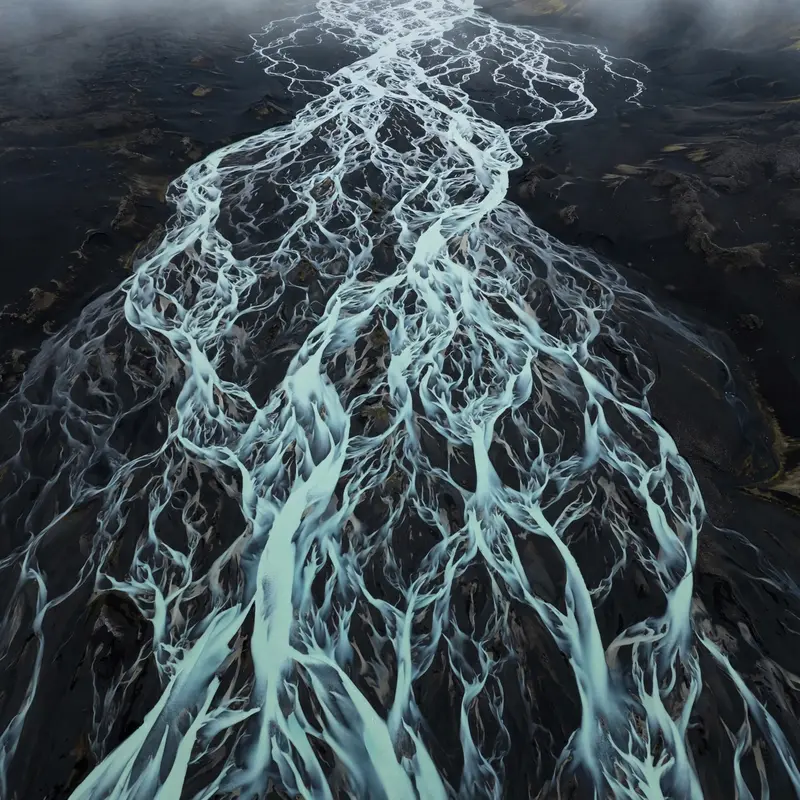 Milky turquoise glacial rivers braiding across a black volcanic plain in Iceland, seen from above like veins. Aerial abstract photography, overcast