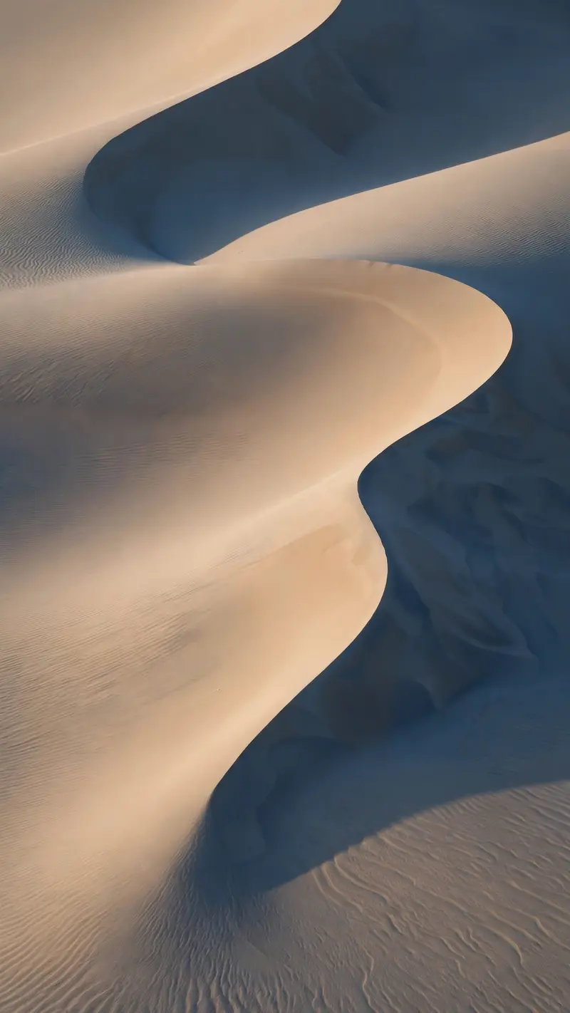 Smooth wind-carved sand dune curves in warm beige and shadow blue, minimalist abstract landscape, vertical 9:16 composition. Fine art desert